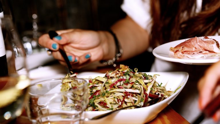 Person with teal nail polish holds dish of prosciutto and serves themselves salad at restaurant