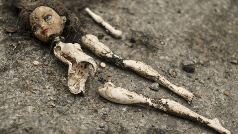 A children's fashion doll, unclothed and broken into pieces, lays on the ground covered in dirt