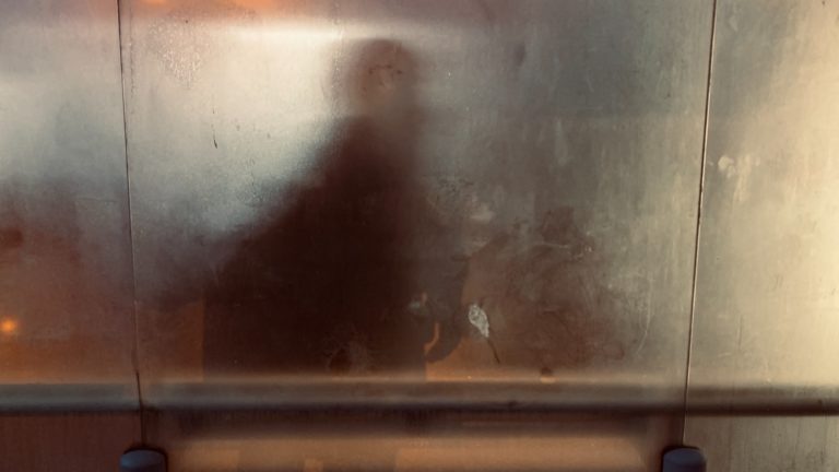 A person looks at their blurred reflection in a dirty metal surface